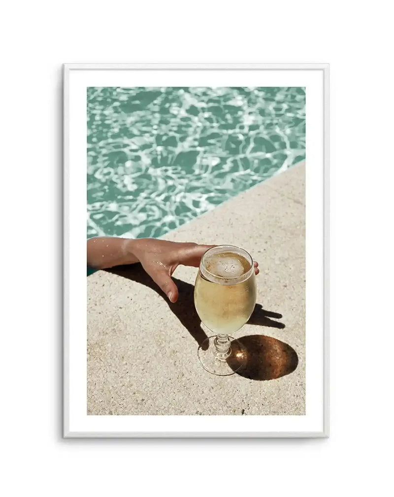 Framed photograph of a hand reaching into a swimming pool next to a glass of white wine, with a white frame and white border. The composition captures the vibrant turquoise water of the pool with rippling light reflections, a tanned hand with water droplets, and a stemmed glass of pale yellow wine casting a long shadow on the light-coloured concrete pool deck.
