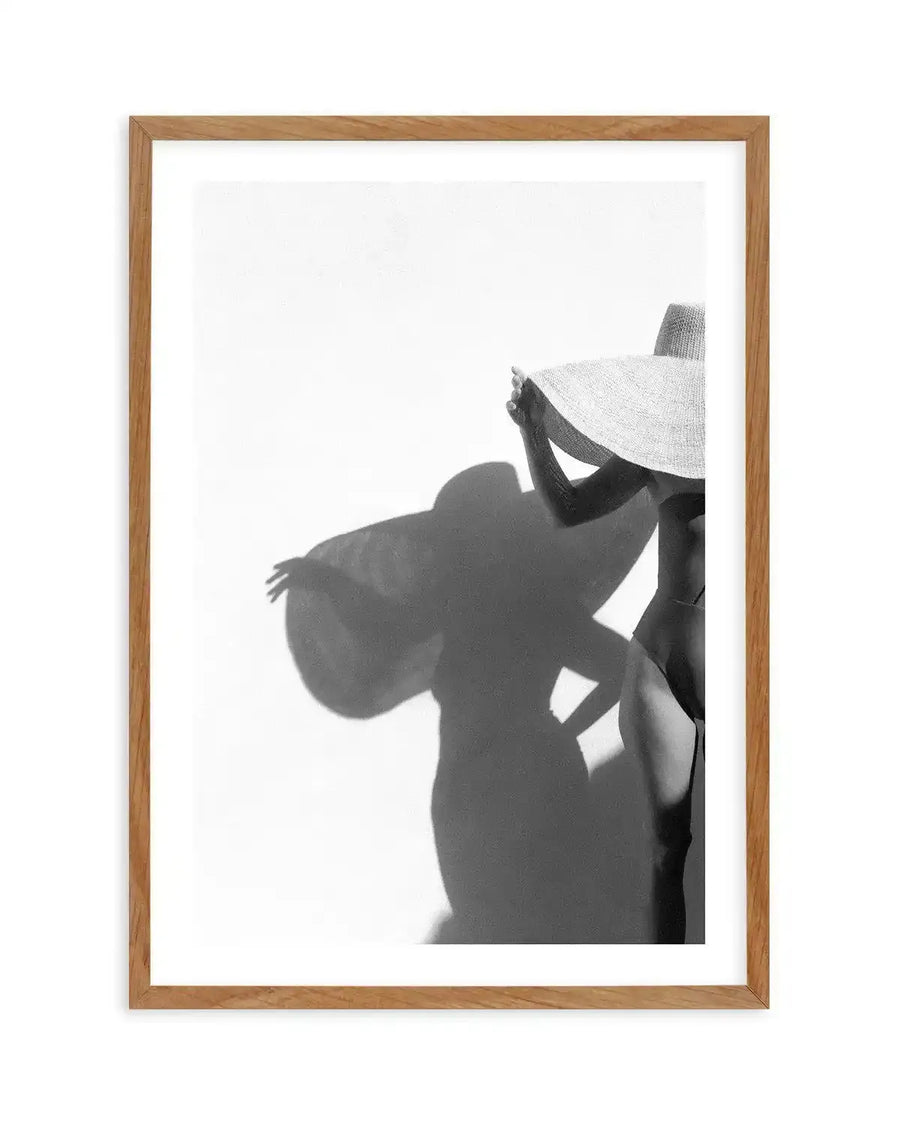 Black and white art print in natural wood frame with white border, featuring a silhouetted figure wearing a wide-brimmed hat, shown from shoulders up against a bright white background. The figure's shadow is cast prominently to the left, creating a minimalist, high-contrast composition with a contemplative mood. Photograph-style artwork with grainy texture.