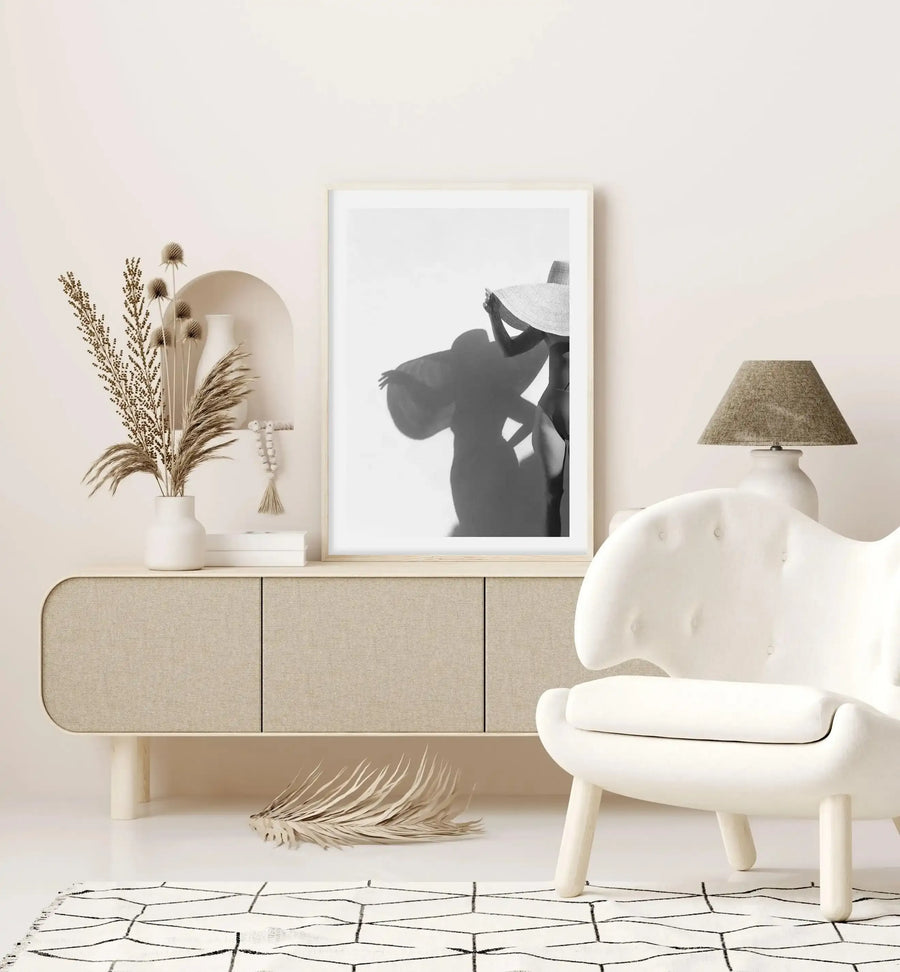 Framed black and white art print displayed on a beige linen credenza in a minimalist room setting. The photograph shows a silhouetted figure wearing a wide-brimmed hat and loose clothing, captured in profile against a white background. The print has a natural wood frame with white border. Styled alongside dried pampas grass in a white ceramic vase, a woven lampshade, and a cream-colored sculptural chair on a geometric patterned rug.