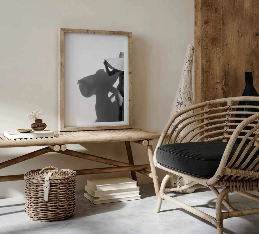 Black and white silhouette art print featuring a woman in profile tilted back with head thrown back and hand near her face, displayed in a natural wood frame with white border on a light wooden desk. The minimalist illustration conveys movement and emotion against a soft white background. The framed print is styled in a bright, airy room setting alongside natural woven furniture, a woven basket, small potted flowers, stacked books, and a dark wine bottle, creating a calm, contemporary aesthetic.