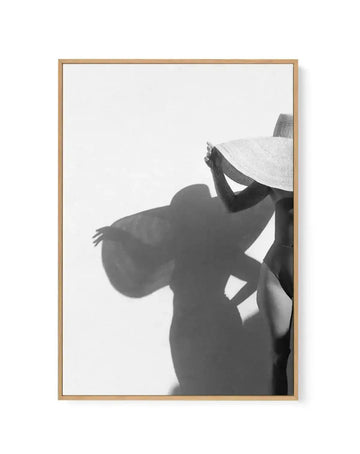 Framed black and white photograph of a woman in a wide-brimmed straw hat and swimsuit, casting a large shadow on a white wall. The woman is partially visible on the right, holding the hat to shield her face, with her shadow prominently displayed on the left, showing her full figure and outstretched arm. The print is displayed in a natural oak frame with a white border.