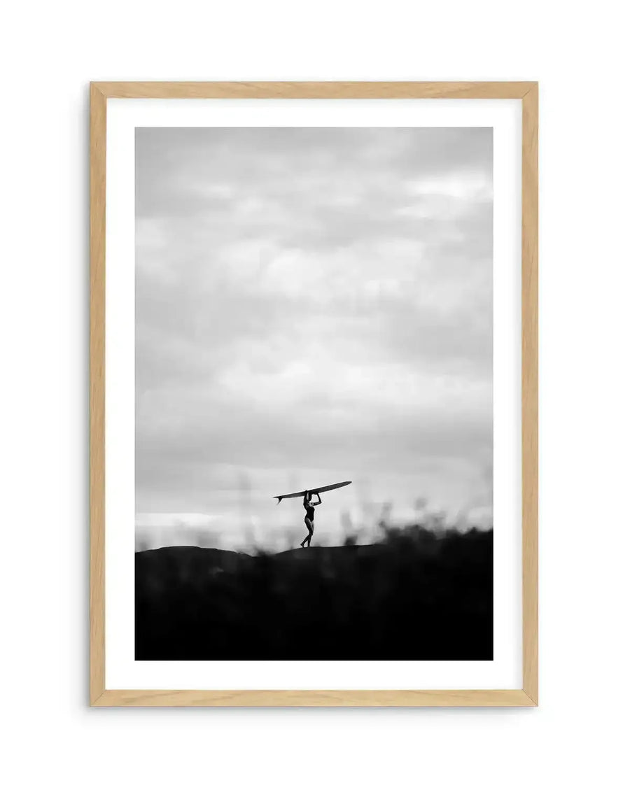 Black and white art print titled "Summer Storm I" by Mario Stefanelli, displayed in a natural wood frame with white border. The photograph shows a silhouetted figure standing on a dark hillside, holding a surfboard overhead against an expansive, cloudy grey sky. The composition uses high contrast between the dark foreground landscape and the dramatic, overcast sky above, creating a moody, minimalist aesthetic.