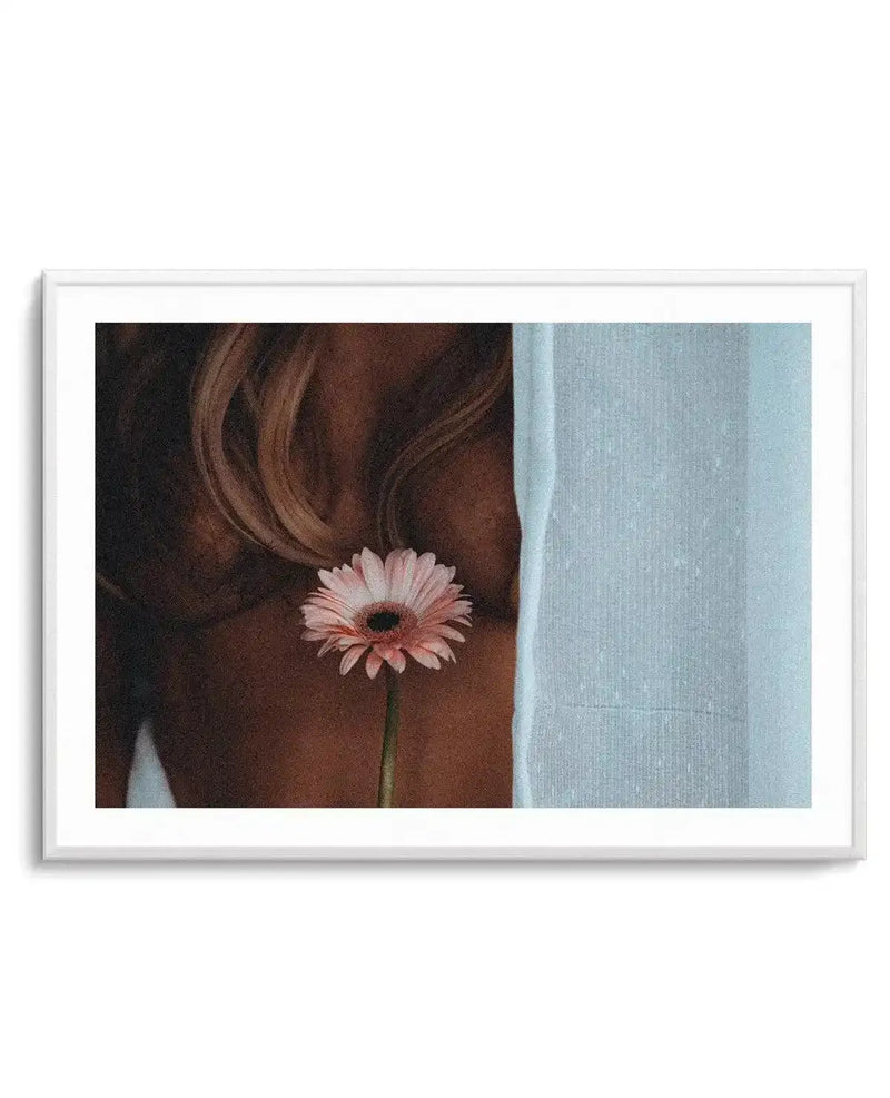 Framed photograph of a person with light brown hair and tanned skin holding a single pink gerbera daisy with a dark center, against a light blue, textured fabric background, with a white frame and white border.