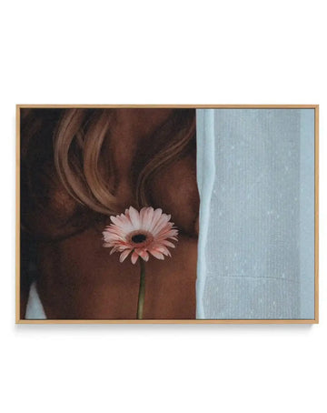 Framed canvas art print titled "Sunday" by Mario Stefanelli, featuring a close-up portrait of a person with long, wavy chestnut brown hair holding a delicate pink gerbera daisy with a dark center. The figure is positioned against a soft blue-grey wall on the right side. The artwork has a warm, intimate quality with rich brown tones contrasting against the cool pale blue background. The canvas is mounted in a natural wood frame with a white border.
