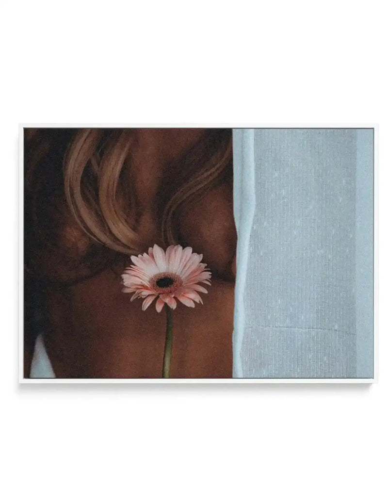 Framed canvas art print titled "Sunday" by Mario Stefanelli. The artwork features a person with long, wavy auburn hair viewed from behind, holding a delicate pink gerbera daisy with a dark center near their shoulder. The left side shows the figure against warm brown tones, while the right side transitions to a cool pale blue-gray textured wall. The composition has a soft, intimate mood with natural lighting. The canvas is mounted in a black frame with a white border.