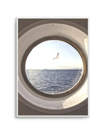 Framed photograph of a seagull flying over the ocean, viewed through a ship's porthole, with a white frame and white border. The circular porthole frames a bright blue sky with a single white seagull in flight, and the deep blue ocean with gentle waves below. Distant land is visible on the horizon under a soft, bright sky.