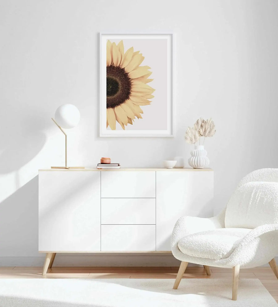 Lifestyle shot of a minimalist living room featuring a framed art print of a sunflower displayed above a white sideboard. The print features a close-up, cropped view of a sunflower with bright yellow petals and a dark brown seed head, set against a light grey background, with a white frame and white border. The room includes a white textured armchair, a white globular lamp with a gold base, and dried palm leaves in a white ribbed vase.