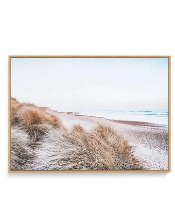 Framed canvas art print featuring a serene coastal landscape photograph. Golden dune grasses in soft focus occupy the foreground on the left side, their dried tan and beige tones textured and windswept. The middle ground shows a wide sandy beach in warm beige tones extending toward calm waters. The ocean stretches across the background in soft blues and grays with gentle waves. The sky is pale, overcast, and misty. The natural wood frame is light tan with a subtle grain, displayed against a white wall.