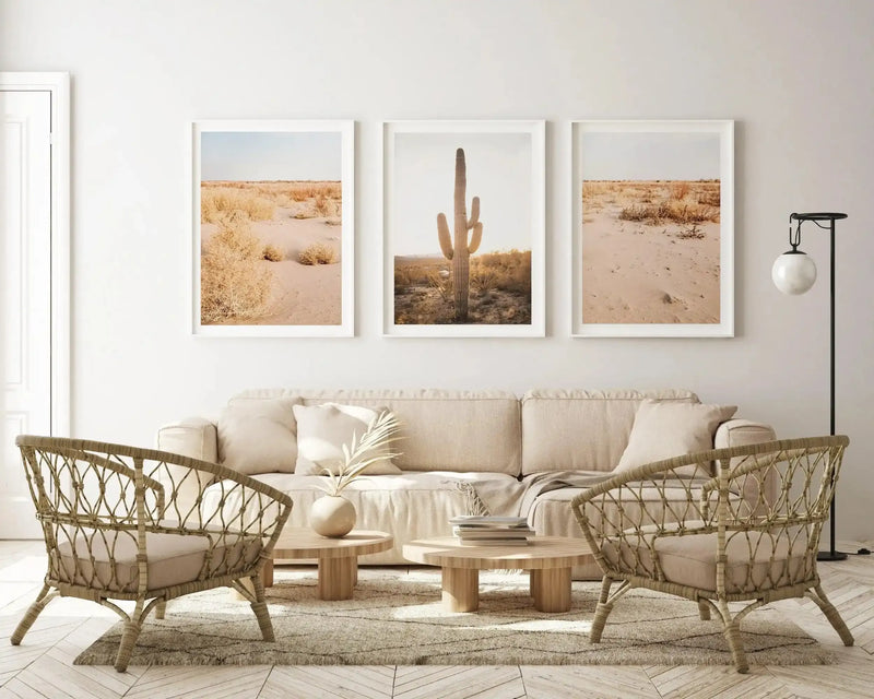 Lifestyle shot of three framed art prints displayed above a cream linen sofa in a living room with a neutral desert aesthetic. The three prints feature desert landscapes with sandy ground and sparse, dry vegetation under a pale blue sky. The central print shows a tall saguaro cactus in the foreground. The room includes two woven rattan armchairs, two round wooden coffee tables, a textured cream rug, and a black floor lamp with a white globe shade.