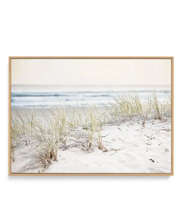 Framed canvas art print featuring a serene beach scene with pale sand dunes and wispy beach grass in soft greens and tans in the foreground. Beyond the dunes, calm ocean waves in muted blues and grays stretch across the middle ground under an overcast, pale sky. The photograph has a soft, muted color palette dominated by whites, creams, and cool tones. The canvas is mounted in a natural wood frame with a white border, creating a calm, coastal aesthetic.