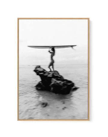 Framed black and white photograph of a lone surfer standing on a rock in the ocean, holding a surfboard over their head, with a natural wood frame and white border. The image has a blurred, motion-like effect, capturing the movement of the surfer and the water.