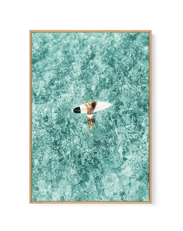 Framed canvas art print featuring an aerial photograph of a shirtless surfer holding a white surfboard, lying on shallow turquoise water. The water displays intricate ripple patterns and varying shades of aqua, teal, and seafoam green. Natural wooden frame with white border. Vibrant, tropical color palette creates a serene coastal aesthetic.