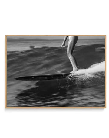 Framed black and white photograph of a surfer riding a wave in Malibu, displayed in a natural oak frame with white border. The composition captures the surfer's lower body and board as they glide across the water, creating a spray of white foam. The ocean stretches to the horizon under an overcast sky, with a sense of motion conveyed by the blurred water.