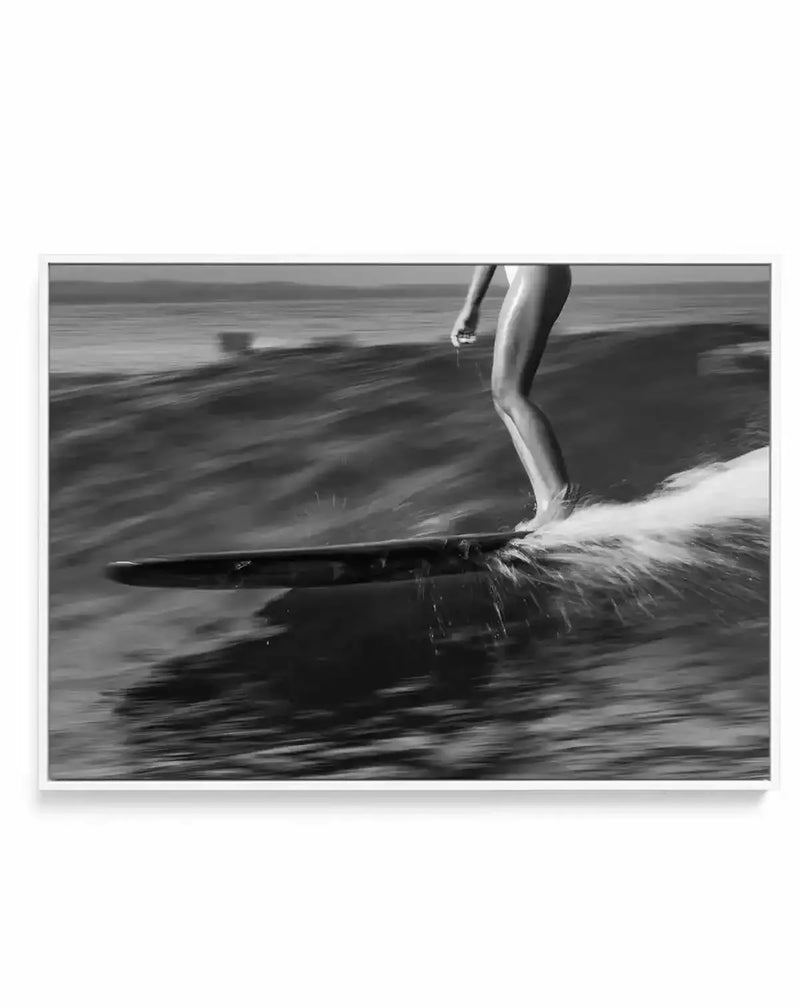 Framed black and white photograph of a surfer riding a wave, printed on canvas and displayed in a white frame. The composition captures the surfer's lower body and legs in motion, with water splashing around the surfboard as it cuts through the wave. The background shows the ocean and horizon, blurred to emphasize movement.