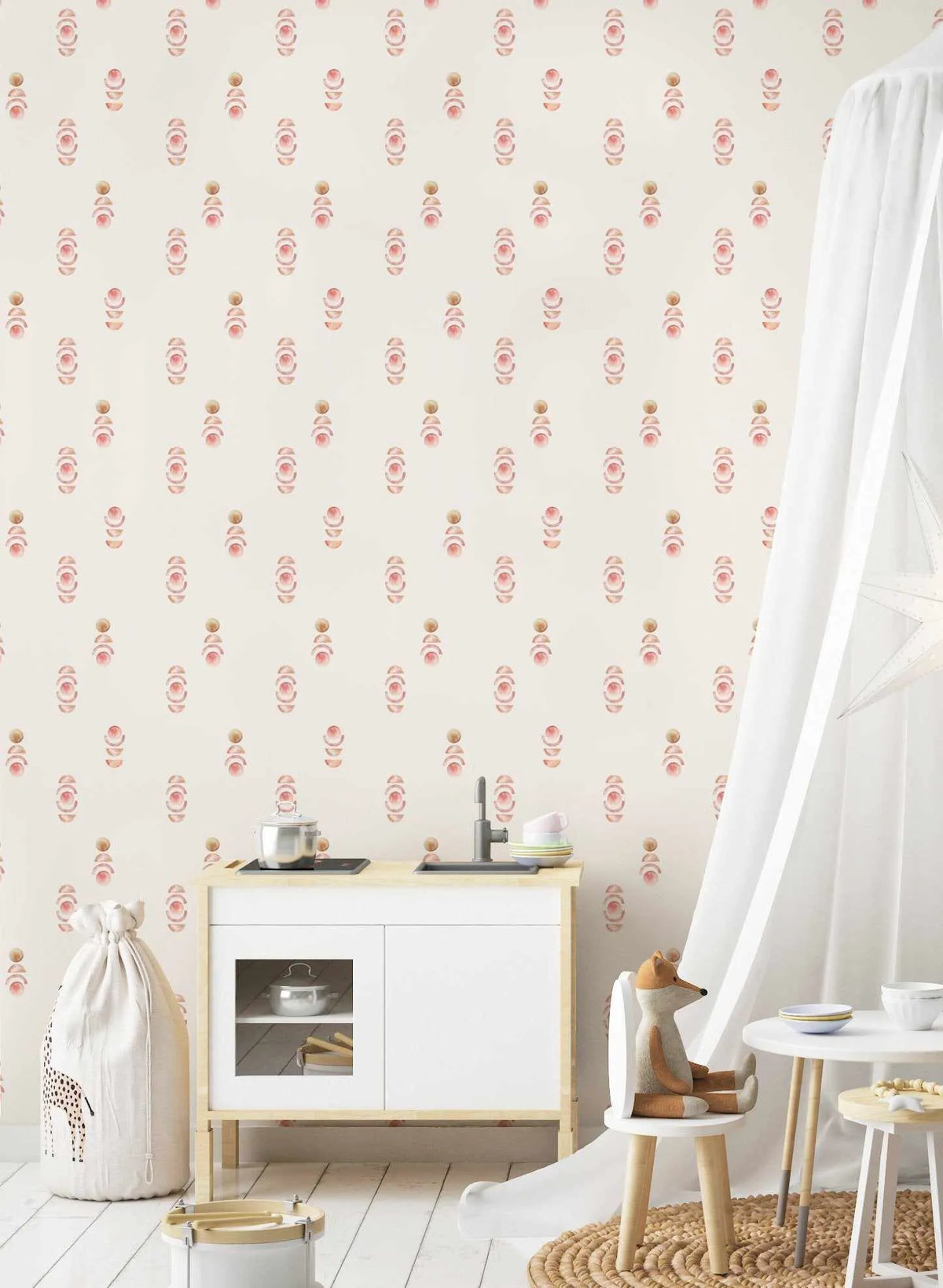 Sweet Boho Wallpaper displayed on a bedroom wall featuring a repeating pattern of hand-painted watercolour designs in dusty rose pink and warm terracotta tones on a cream background. The motif shows stacked circular and oval shapes with natural wood bead accents, creating a boho aesthetic. The wallpaper is shown in a styled children's room setting with natural wood furniture, white accents, and woven textures, demonstrating the pattern's soft, calming appearance in an interior space.