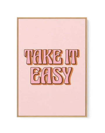 Framed canvas art print featuring the text "TAKE IT EASY" in large, bold, retro-style lettering with orange and brown gradient shading and dark burgundy outlines. The text is centered on a soft blush pink background. The canvas is surrounded by a natural wood-tone frame with a white border, creating a clean, minimalist presentation against a white background.