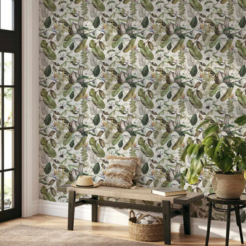 Lifestyle shot of a room with Tathra wallpaper, featuring a repeating pattern of lush green tropical foliage and white bird of paradise flowers on a light background. The wallpaper covers a wall next to a black-framed glass door, with a woven bench, a straw hat, a textured cushion, and a potted plant in the foreground.