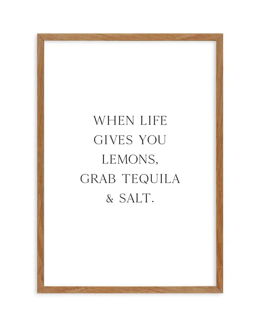 Framed art print with natural wood frame displaying black serif text on white background that reads "WHEN LIFE GIVES YOU LEMONS, GRAB TEQUILA & SALT." The text is centered and evenly spaced across the white interior, creating a minimalist, typographic design with a humorous tone.
