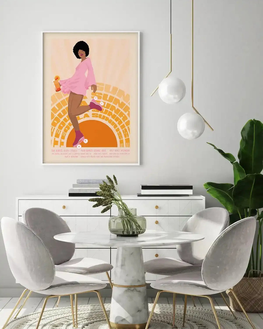 Lifestyle shot of a dining area featuring a framed art print displayed above a white console table. The print is an illustration of a woman with an afro, wearing a pink dress and roller skates, holding a drink, against a background of a setting sun with text "Tequila Sunrise". The room includes a round marble table with four grey velvet chairs, two pendant lights with white spherical shades, and a large green plant in a woven basket.