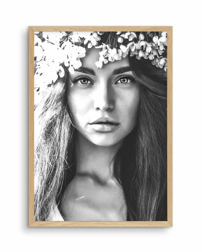 Black and white photographic art print in a light natural wood frame featuring a close-up portrait of a woman with long straight hair and intense gaze, wearing a white flowering crown of delicate clustered blooms across the top of her head. The high-contrast monochromatic photograph emphasizes fine facial details and hair texture against a soft white background. The artwork is matted with a white border within the wooden frame.