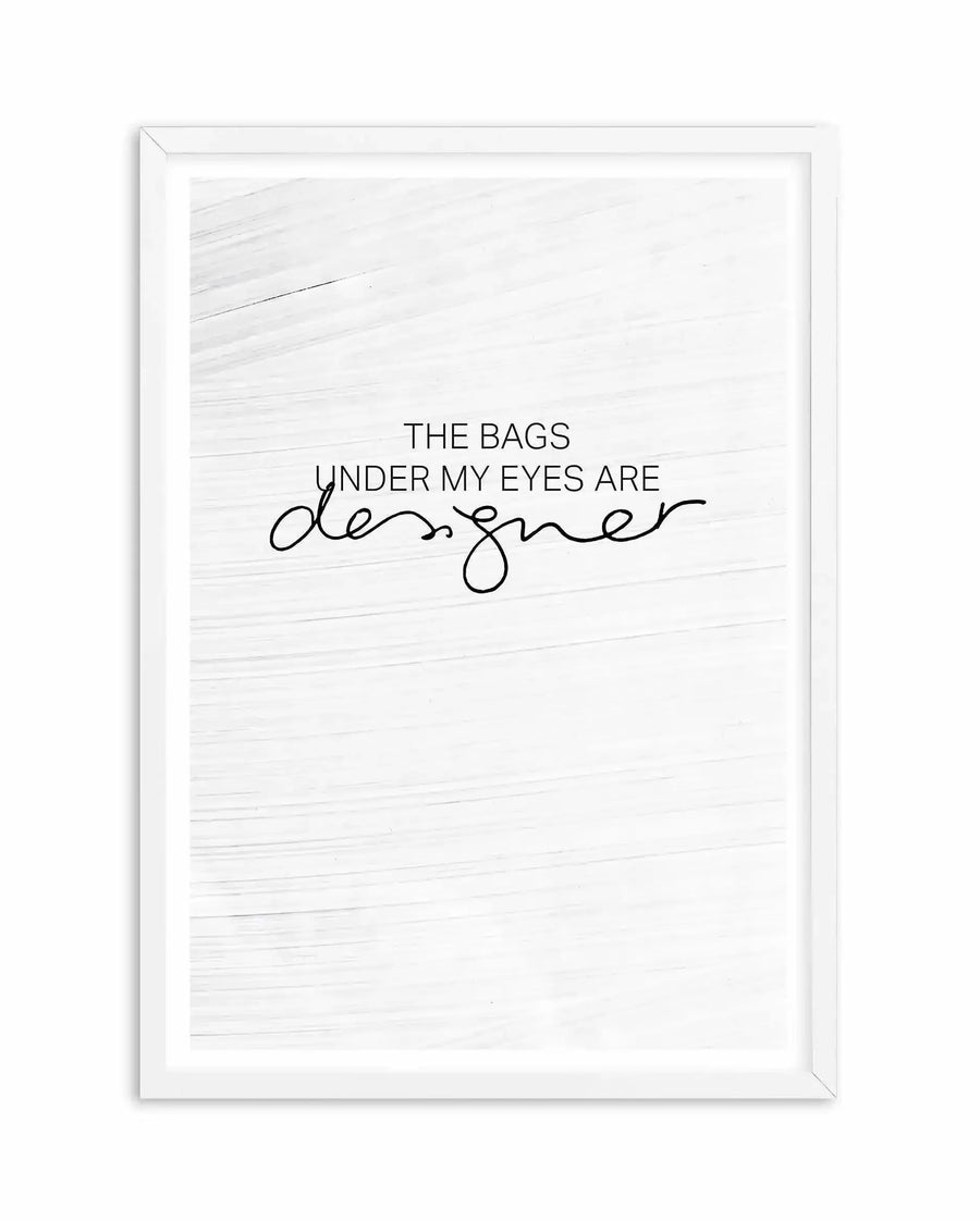 White framed art print featuring a minimalist design on a white textured background. The text reads "THE BAGS UNDER MY EYES ARE" in sans-serif font, with "designer" written in an elegant, flowing cursive script below. The frame is white wood with a white border surrounding the artwork.
