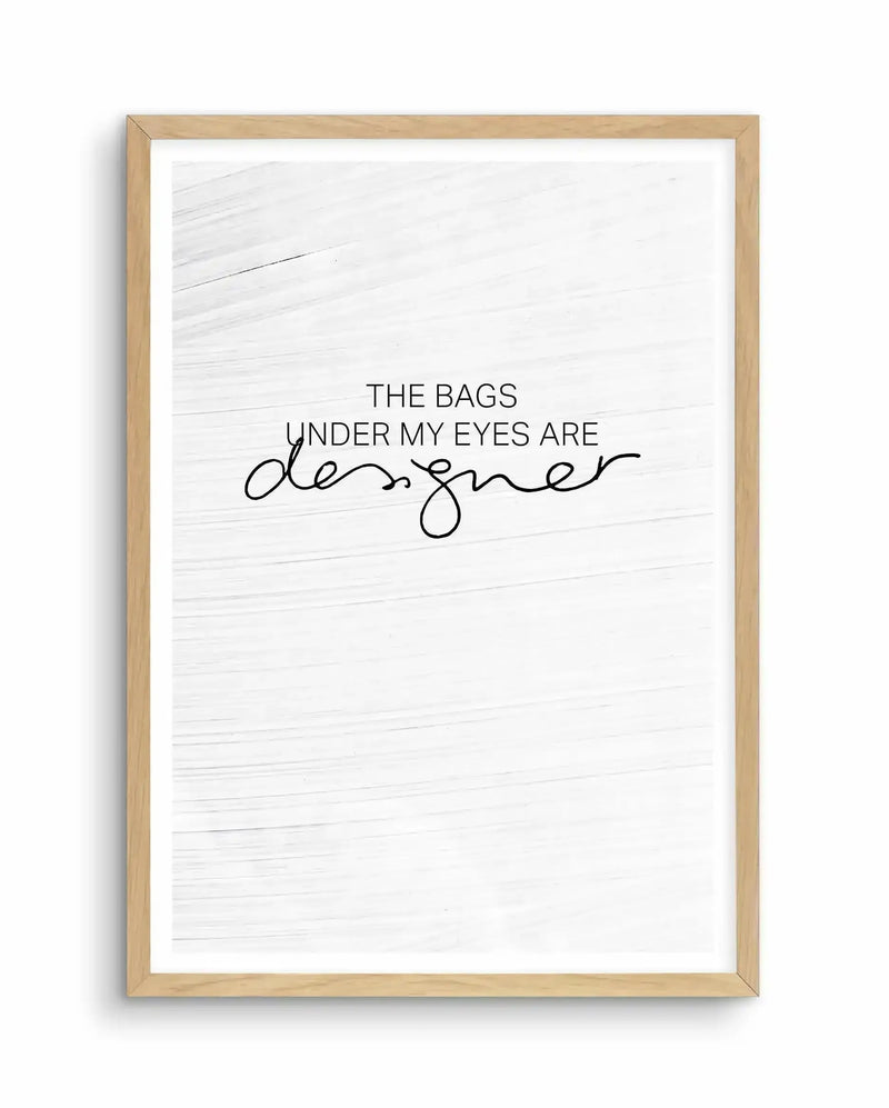 Framed art print with natural wood frame containing white textured background. Text reads "THE BAGS UNDER MY EYES ARE" in sans-serif font, with "designer" written below in flowing cursive script. Minimalist design with black text on off-white background with horizontal wood grain texture visible throughout.