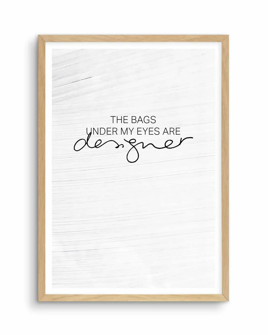 Framed art print with natural wood frame containing white textured background. Text reads "THE BAGS UNDER MY EYES ARE" in sans-serif font, with "designer" written below in flowing cursive script. Minimalist design with black text on off-white background with horizontal wood grain texture visible throughout.