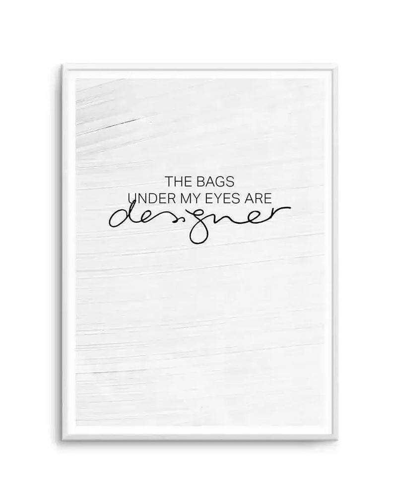 Framed art print featuring white shiplap wood texture background. Text reads "THE BAGS UNDER MY EYES ARE" in sans-serif font, with "designer" written in flowing black script handwriting below. White frame with white border. Minimalist, humorous design in black and white.