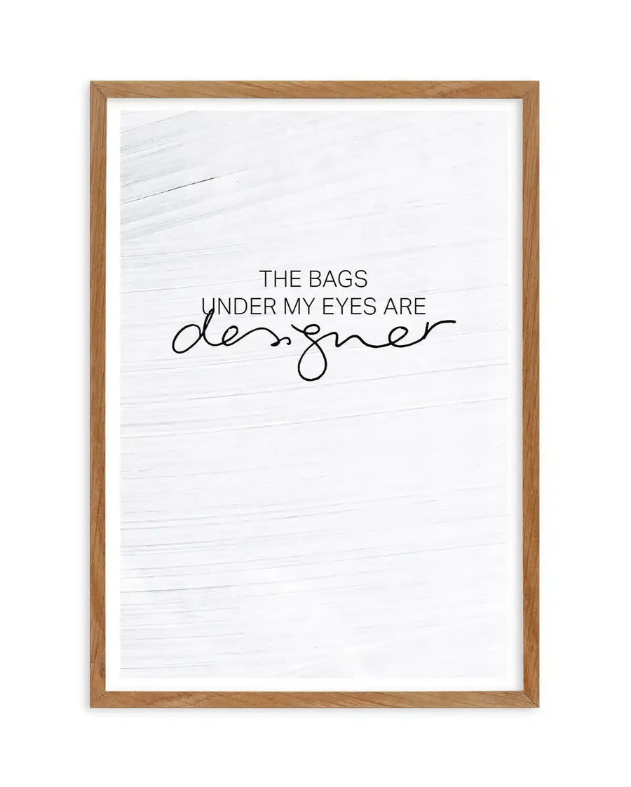 Framed art print with light wood frame displaying text on a white shiplap-textured background. The design reads "THE BAGS UNDER MY EYES ARE" in sans-serif capital letters, with "designer" written below in a flowing, continuous cursive script. Black text on off-white background with horizontal wood grain texture visible throughout. White border surrounding the artwork within the frame.