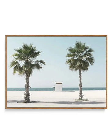 Framed canvas art print featuring a minimalist beach scene with two tall palm trees flanking a white lifeguard tower, set against calm turquoise ocean and pale sand under a clear light blue sky. Natural wood frame with horizontal orientation. Photograph-style imagery with soft, muted colour palette and serene coastal atmosphere.