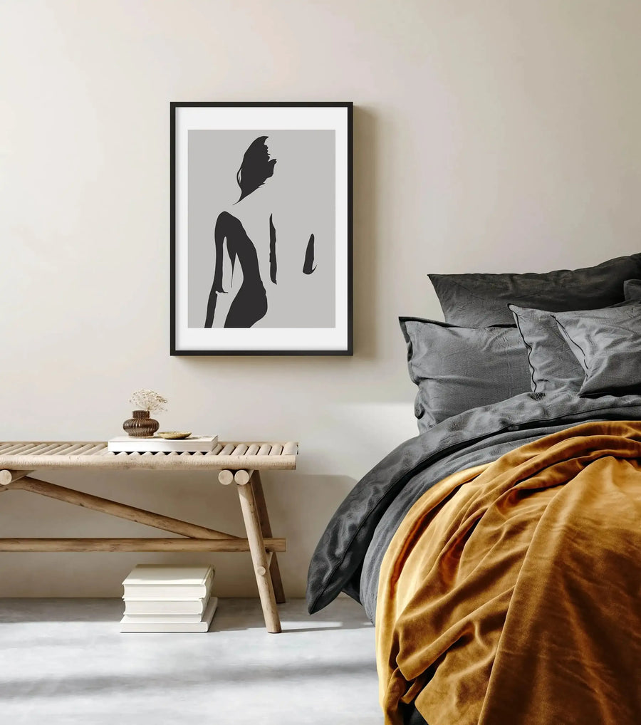 Lifestyle shot of a minimalist bedroom featuring a framed art print above a bed. The print features a black silhouette of a woman's back and head against a light grey background, with a black frame and white border. The bed has dark grey pillows and duvet, with a mustard yellow throw blanket. A light wood slatted bench with a small vase of dried flowers and a stack of white books sits beside the bed.
