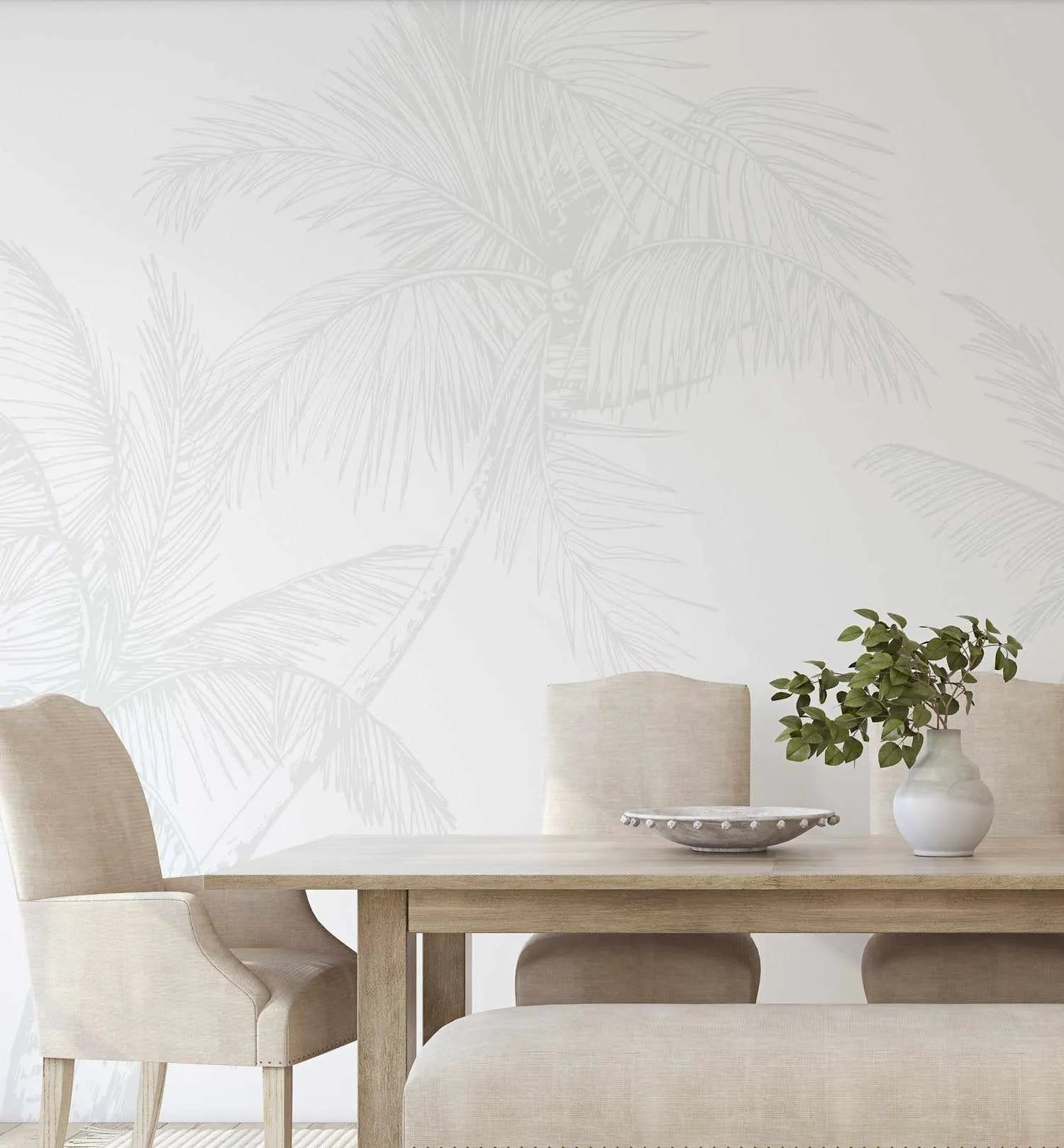 Dining Room:
"The Palms Wallpaper in Grey displayed in a bright dining room with a light wood dining table and upholstered chairs. The wallpaper features a repeating pattern of large-scale palm tree fronds and trunks in a soft, muted grey against a clean white background, creating a subtle tropical motif. A white ceramic vase with green foliage and a decorative bowl sit on the table."