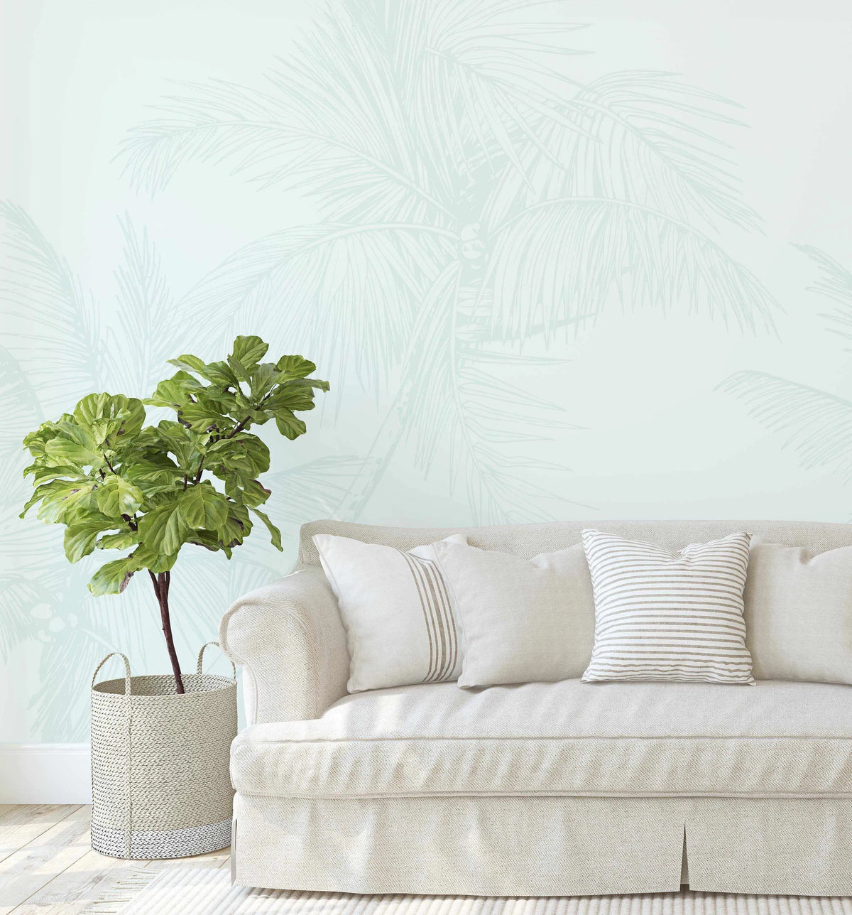 Lifestyle shot of a living room with The Palms Wallpaper in Seafoam. The wallpaper features a repeating pattern of palm fronds in a subtle, light seafoam green on a slightly lighter seafoam background, creating a soft, tonal effect. A cream-colored sofa with throw pillows in cream and striped cream and beige is positioned in front of the wallpaper. To the left, a large fiddle-leaf fig plant in a woven basket sits on a light wood floor with a cream rug.