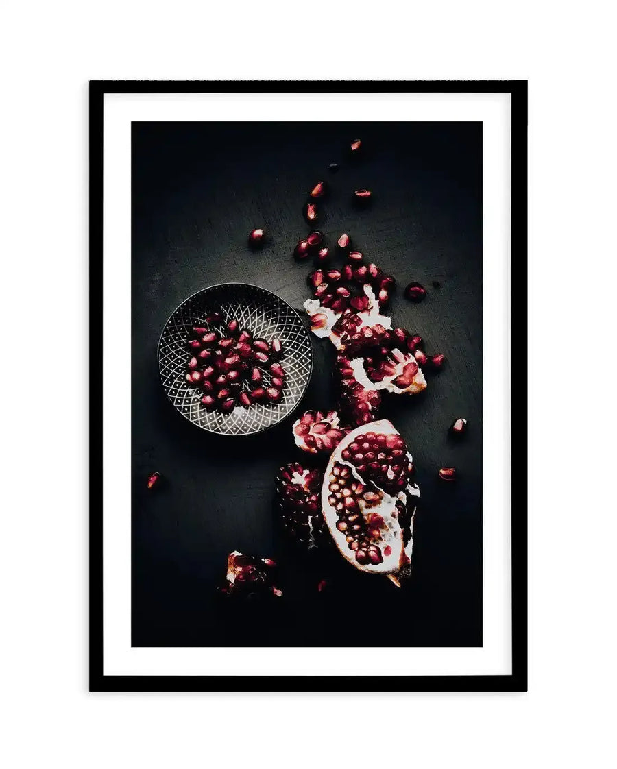 Framed photograph of scattered pomegranate seeds and fruit on a dark surface, with a black frame and white border. A small, dark grey bowl with a geometric pattern holds a pile of seeds in the upper left, while a broken pomegranate and loose seeds are spread across the lower right, revealing the deep red arils and white pith against the dark background.