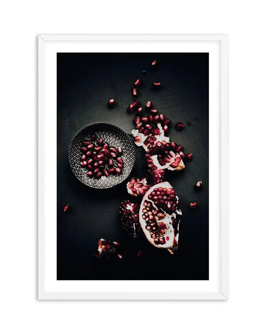 "Framed photography art print featuring a close-up, overhead view of a deconstructed pomegranate on a dark, almost black background, with a white frame and white border. The composition shows a small, dark grey bowl with a geometric pattern filled with bright red pomegranate seeds on the left, and a broken pomegranate with scattered seeds and pieces of white pith on the right. The rich red seeds contrast with the dark background, creating a dramatic and moody aesthetic."