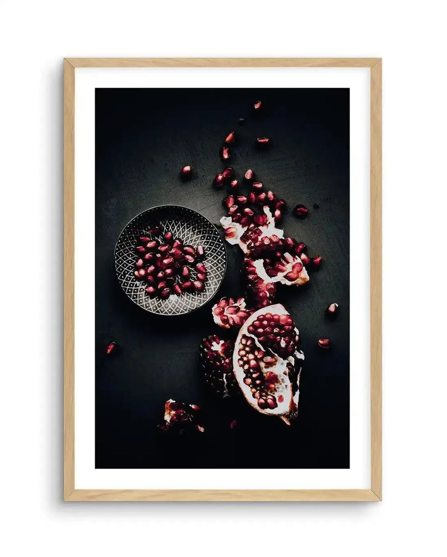 "Framed photography art print of pomegranates on a dark background, displayed in a natural oak frame with white border. The composition features a halved pomegranate and scattered seeds, with a small patterned bowl of seeds on the left, all against a deep charcoal surface."