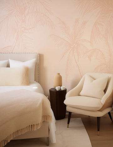Soft terracotta pink wallpaper mural featuring delicate palm frond illustrations in a botanical line-drawing style, displayed in a serene bedroom setting with a cream upholstered bed, matching armchair, dark wood side table with ceramic cups, and neutral bedding with fringe trim.
