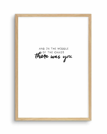 Art print in natural wood frame featuring centered black hand-lettered text on white background. Upper portion reads "AND IN THE MIDDLE OF THE CHAOS" in thin sans-serif font. Below, "there was you" appears in flowing, connected cursive script. Minimalist design with ample white space, creating contemplative mood. Frame has warm honey-toned wood finish with subtle grain visible.