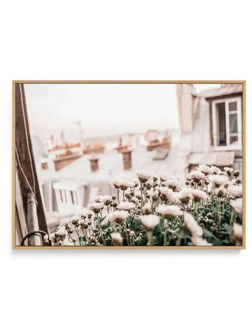 Framed canvas art print featuring soft-focus photograph of white carnation flowers in foreground with green stems, overlooking a Parisian rooftop with cream-colored buildings, chimneys, and a window visible on right side. Image has warm, muted color palette with dusty rose and beige tones creating dreamy, vintage aesthetic. Natural wooden frame with white border around photograph.
