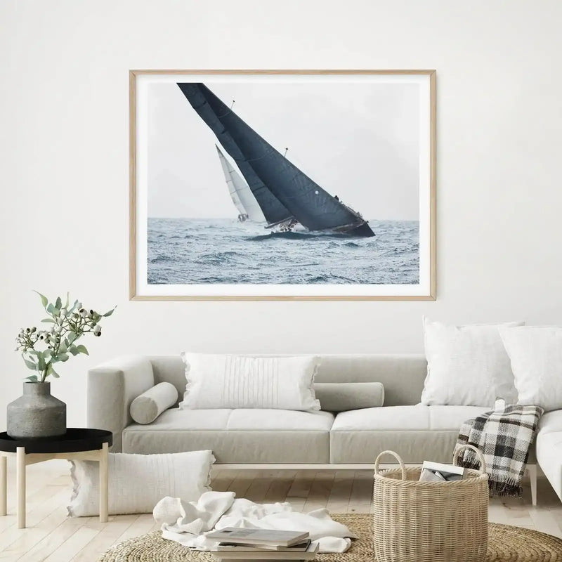 Lifestyle shot of a minimalist living room featuring a framed art print above a light grey sofa. The print is a photograph of two sailboats racing on a choppy sea under an overcast sky, with a natural wood frame and white border. The room includes white throw pillows, a woven basket, and a plant in a grey vase on a black side table.