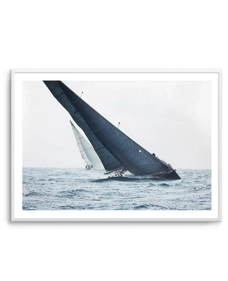 Framed photograph of two sailboats racing on a choppy ocean under an overcast sky, with a large dark blue sail in the foreground leaning heavily to the right, and a smaller white sail in the background. The print is displayed in a white frame with a white border.