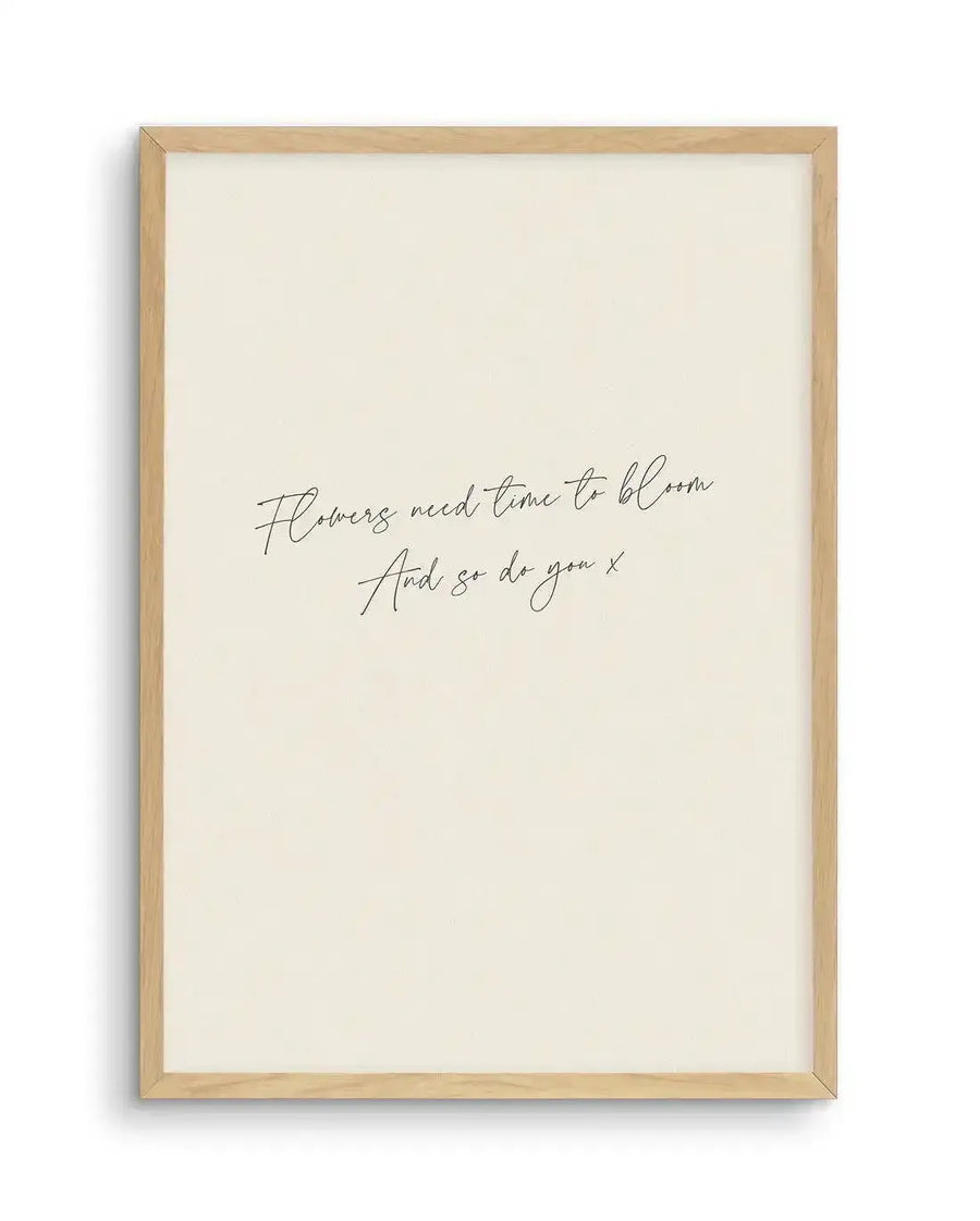Art print featuring the handwritten quote "Flowers need time to bloom And so do you x" in black script on a cream background, displayed in a natural oak frame with white border.