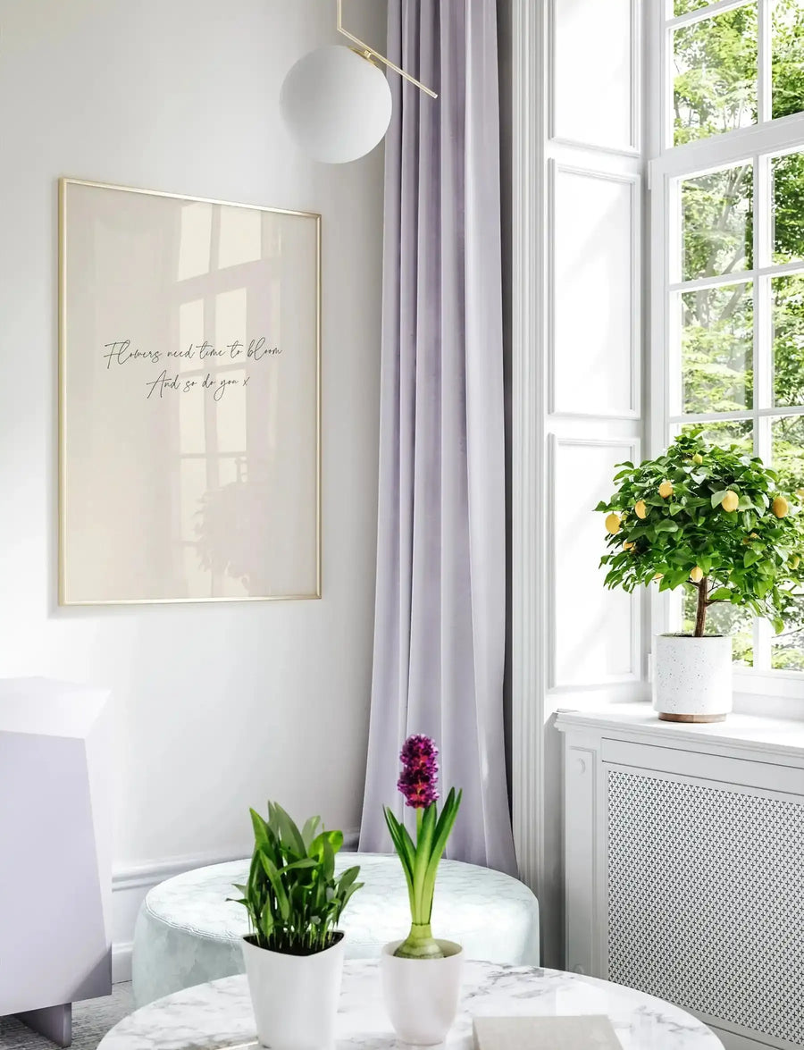 Lifestyle shot of a framed art print displayed on a white wall in a bright, airy room. The print features the handwritten script text "Flowers need time to bloom. And so do you x" in black on a cream background, with a thin gold frame. The room has a large window with white frames and light purple curtains, a potted lemon tree on the windowsill, and two small potted plants on a marble-top table in the foreground.