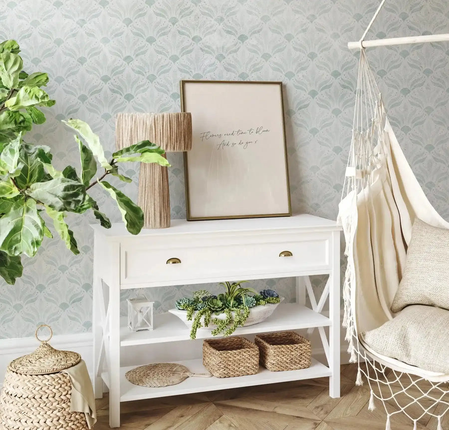 Framed art print displayed on a white console table in a light and airy room with patterned wallpaper. The print features the quote "Flowers need time to bloom. And so do you x" in black script on a cream background, with a thin gold frame. The room includes a large green plant, a woven lamp, a white console table with brass handles, a white hanging chair with cream cushions and throw, and woven baskets. The wallpaper has a repeating pattern of light blue-green and white botanical motifs.