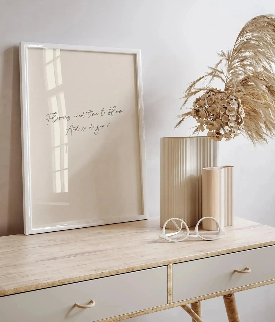 Lifestyle shot of a framed art print on a light wood console table with two drawers. The print features the handwritten quote "Flowers need time to bloom And so do you x" in black script on a cream background, displayed in a white frame with a white border. To the right, two ribbed cream vases hold dried pampas grass and light brown dried flowers. A pair of white, round-framed glasses rests on the table in front of the vases.