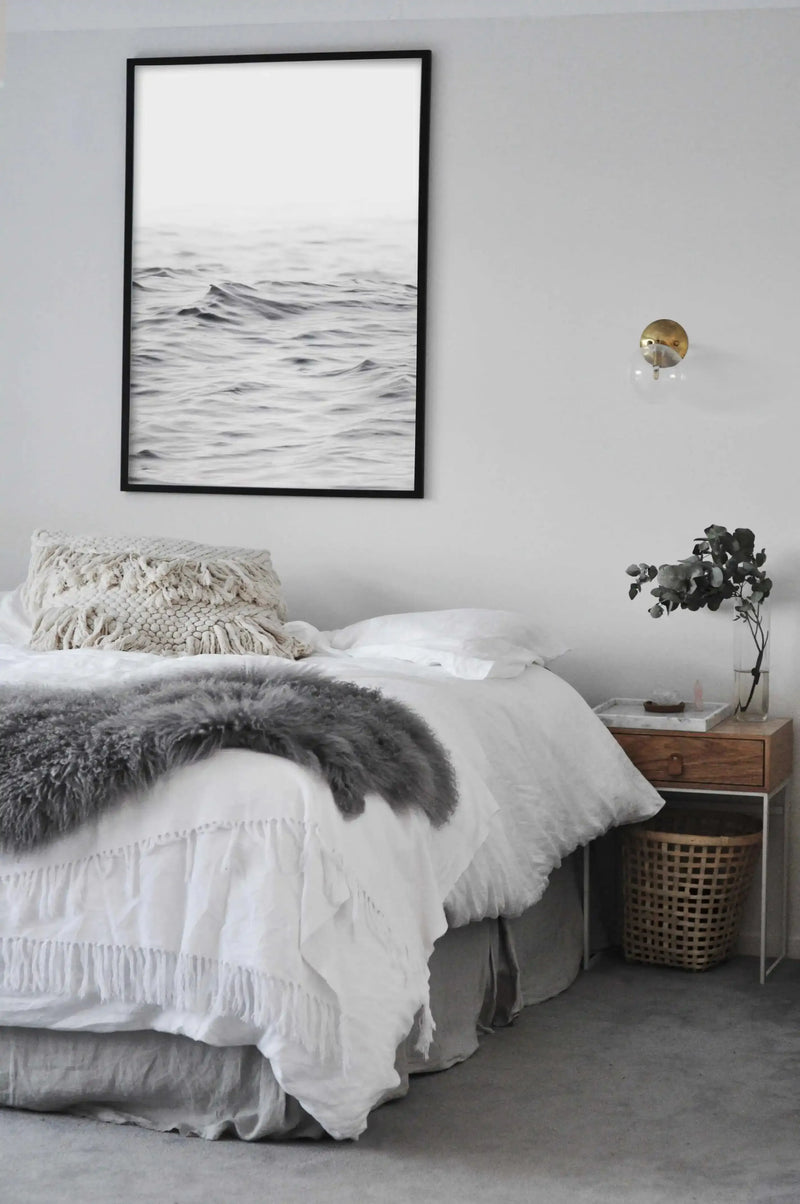 Black-framed art print displaying a minimalist black and white photograph of ocean waves with a pale grey sky, hung above a bed styled with white textured bedding, a cream macramé pillow, grey faux fur throw, and natural wood nightstand with potted eucalyptus and woven storage basket.