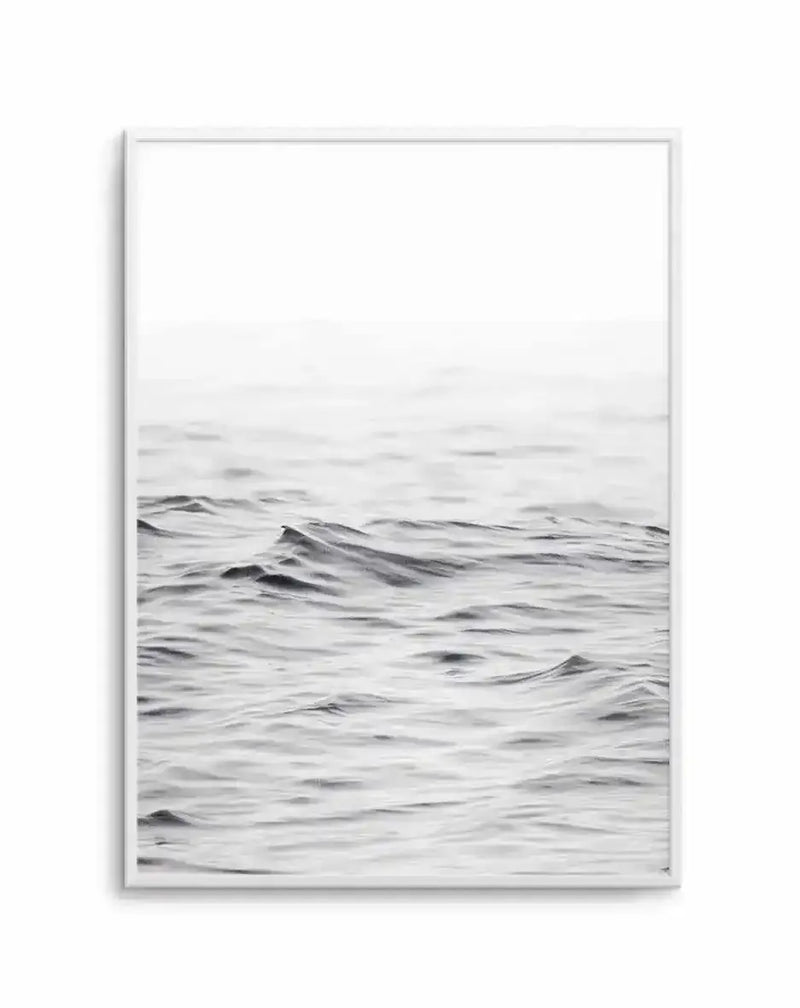 Framed art print displaying a minimalist black and white seascape photograph. The composition shows a calm ocean with gentle rolling waves in varying shades of gray, occupying the lower two-thirds of the frame. The upper portion features an overcast, hazy sky in soft white and light gray tones. The photograph has a serene, muted quality with subtle texture visible in the water's surface. The artwork is presented in a light gray or silver frame with a white border around the image.