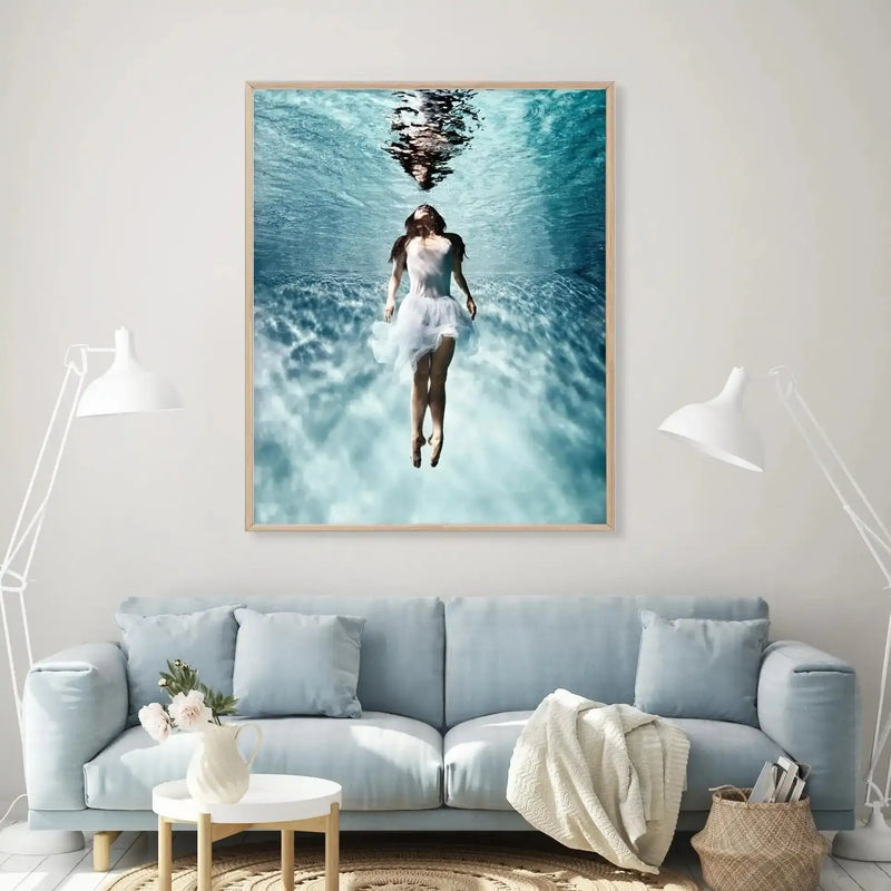 Large framed art print displayed above a light blue sofa in a bright living room. The print features an underwater photograph of a woman in a white dress floating upwards towards the surface, with her reflection visible on the rippling water above. The room includes two white floor lamps, a round white coffee table with a white pitcher and pink flowers, and a woven basket with magazines.