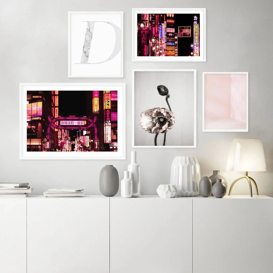 Tokyo Red Light District II Art Print displayed in a white frame with white border, showing a vibrant nighttime photograph of densely packed neon signs in magenta, pink, purple, and yellow hues with Japanese characters, mounted on a light gray wall above a white storage unit styled with ceramic vessels and decorative objects.