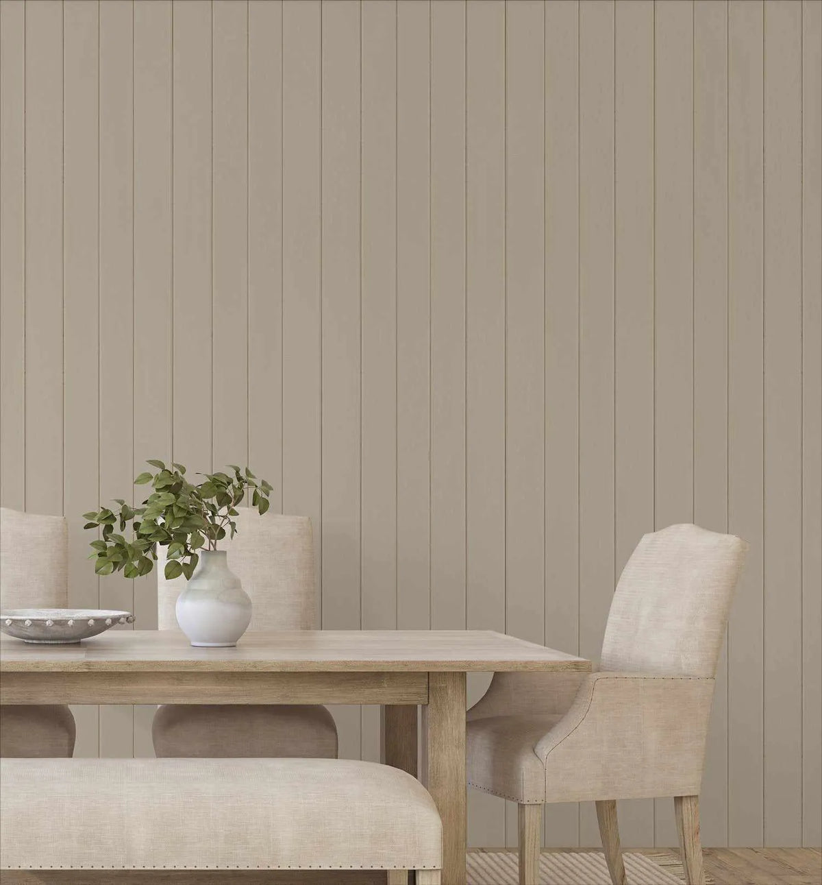 Tongue & Groove Wood Panel Wallpaper in Pebble displayed on a dining room wall behind a light natural wood table. The wallpaper features a vertical shiplap pattern in soft greige with subtle gray undertones, creating the appearance of individual wooden planks. A cream upholstered dining chair sits at the table alongside a white ceramic vase holding fresh green foliage, a metallic bowl, and a cream linen napkin, demonstrating the warm, neutral aesthetic the wallpaper creates in a minimalist interior setting.